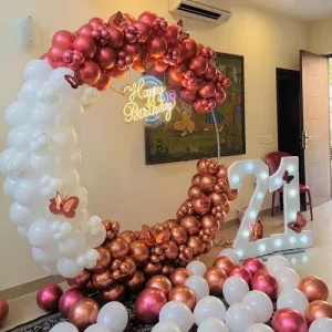 Rose gold and white crescent-shaped balloon arch 21st birthday decoration with butterfly accents and light-up marquee numbers in Indore